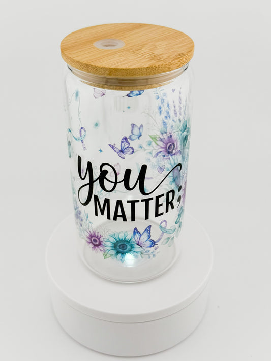 A clear glass tumbler with a light wood bamboo lid, decorated with watercolor-style blue and purple flowers and butterflies. The tumbler features the printed text 'you MATTER' in black script and block lettering.