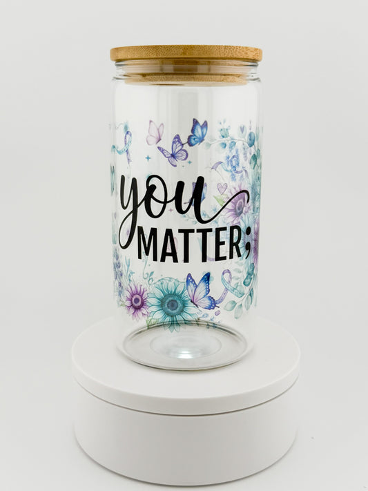 A clear glass jar with a light brown wooden lid, featuring the printed text 'you MATTER' in black script and block lettering, surrounded by blue, purple, and teal butterflies and flowers. The jar is displayed on a round white pedestal against a clean white background.